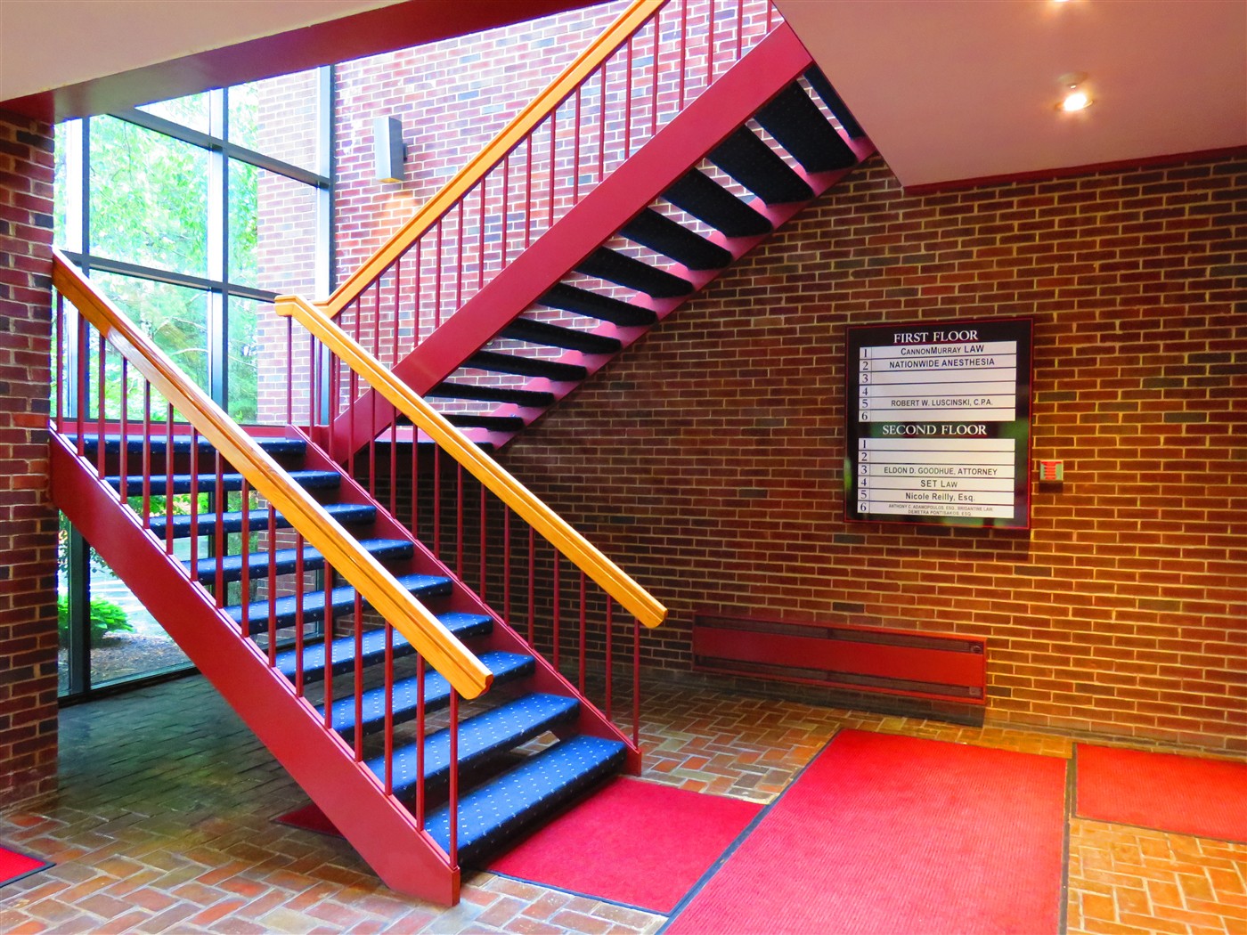 Red staircase with wooden handrails and a directory board on the brick wall.